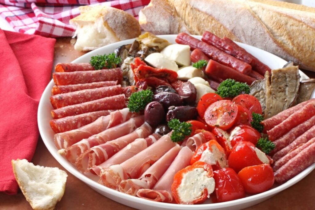 A platter of assorted charcuterie featuring salami, prosciutto, and pepperoni, accompanied by olives, stuffed cherry peppers, mozzarella balls, artichokes, and cherry tomatoes. Slices of baguette are placed around the dish.