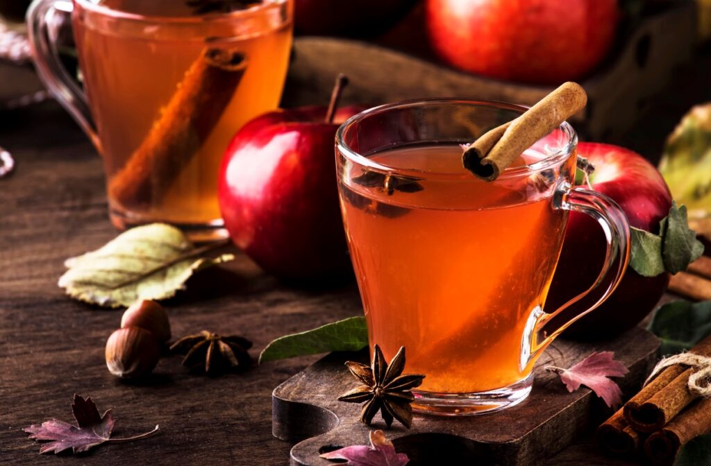 A warm glass mug of apple cider with a cinnamon stick and star anise sits on a wooden table surrounded by fresh apples, autumn leaves, and hazelnuts, creating a cozy fall scene.