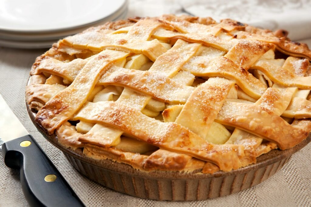 A freshly baked apple pie with a golden brown lattice crust is displayed on a textured surface. The pie is dusted lightly with sugar, and there is a black-handled knife nearby. Plates are stacked in the background.