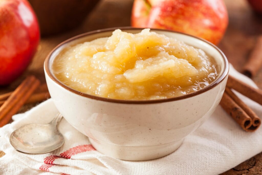 A bowl of applesauce on a cloth napkin with a spoon beside it. Fresh apples and cinnamon sticks are in the background, suggesting flavors of the dish. The setting has a warm and rustic feel.