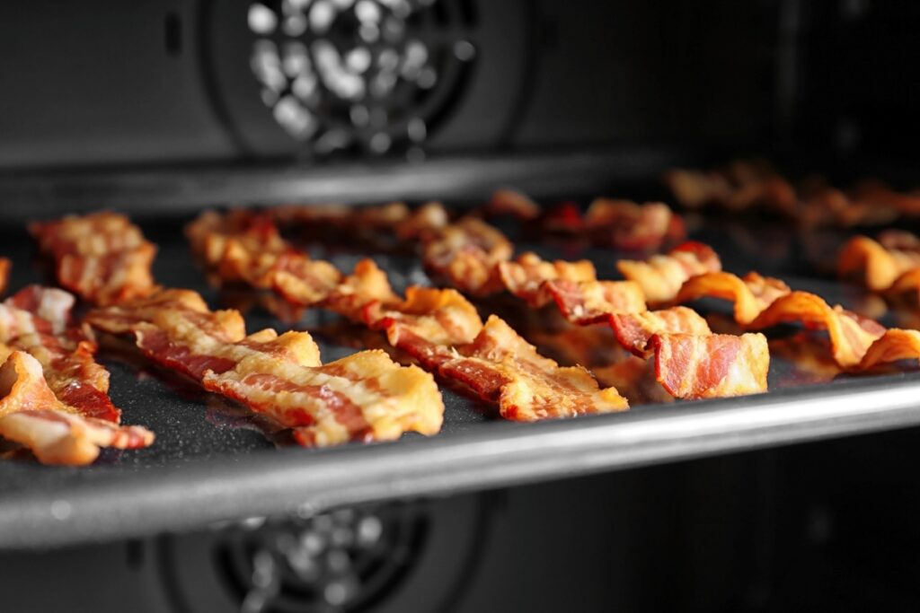 Strips of bacon being cooked in an oven on a tray, with the oven's fan in the background.