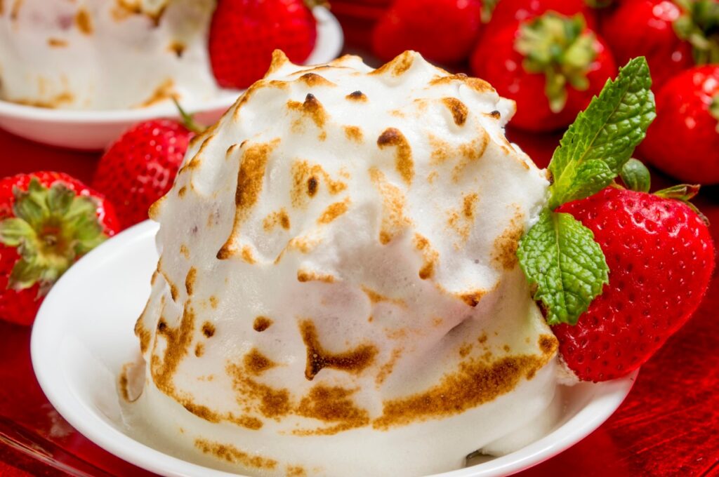 A dessert featuring a golden-brown baked Alaska, topped with fluffy, toasted meringue, sits in a white dish. Fresh strawberries and a sprig of mint decorate the plate, with more strawberries in the background.