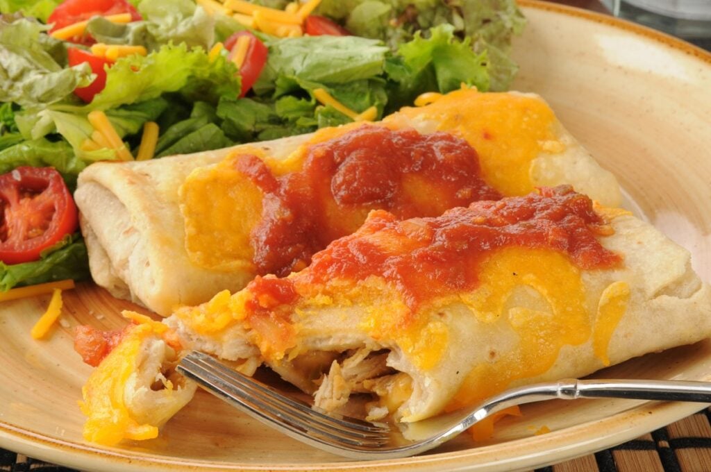 A delicious burrito topped with melted cheese and salsa is partially cut with a fork resting on the plate. A fresh salad with shredded cheese and tomato wedges accompanies the dish, set on a beige plate.