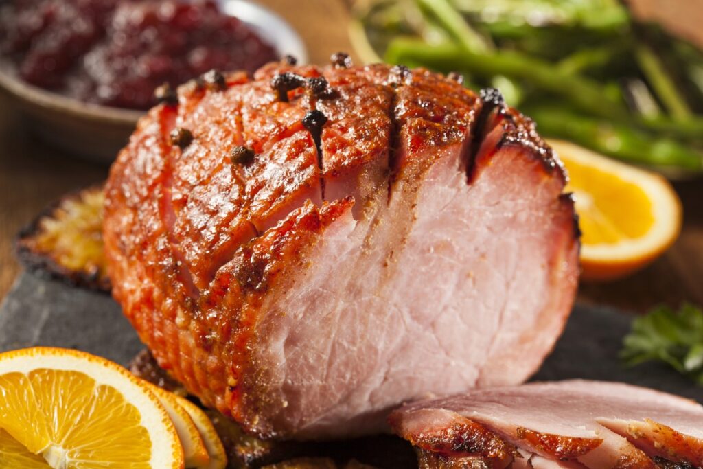 A glazed ham garnished with cloves is sliced and served on a platter. Fresh slices of orange and sprigs of parsley surround the ham. In the background, there’s a bowl of cranberry sauce and cooked green beans.