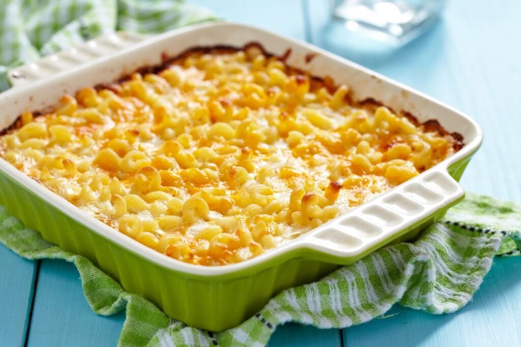 A baked macaroni and cheese dish in a green ceramic baking dish, placed on a green and white checkered cloth on a light blue wooden table. The cheese on top is golden and slightly crispy.