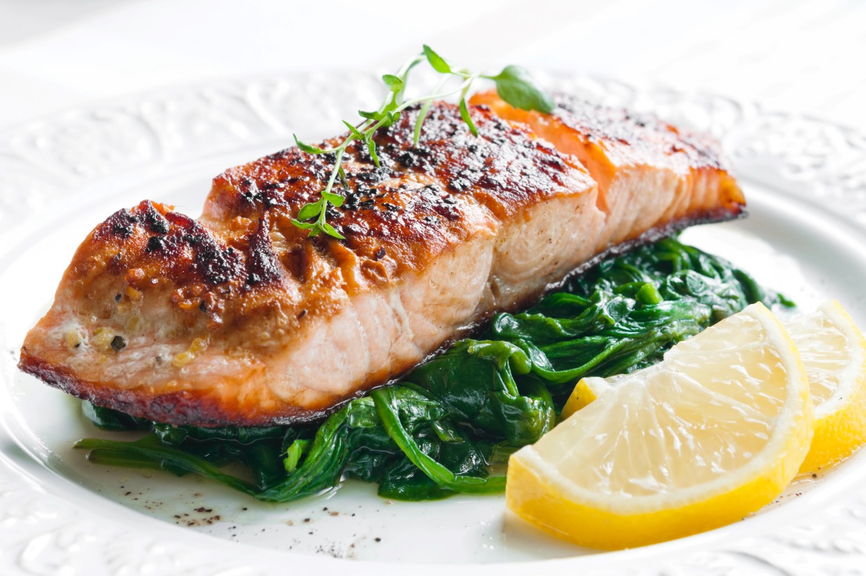 A grilled salmon filet garnished with fresh herbs rests on a bed of sautéed spinach. Two lemon wedges are placed beside the salmon, all served on a white, decorative plate.