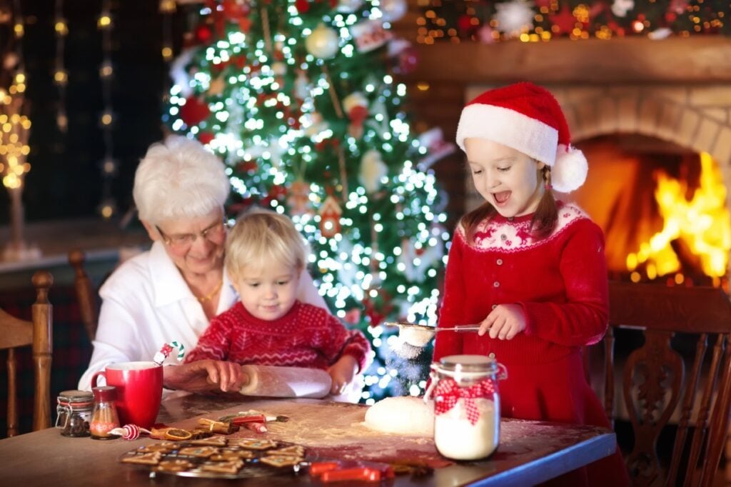 Grandmother and two children bake cookies at a table near a decorated Christmas tree and a lit fireplace. The girl wears a Santa hat and red dress, while the boy has a red sweater. Ingredients and a jar are on the table. Warm, festive atmosphere.