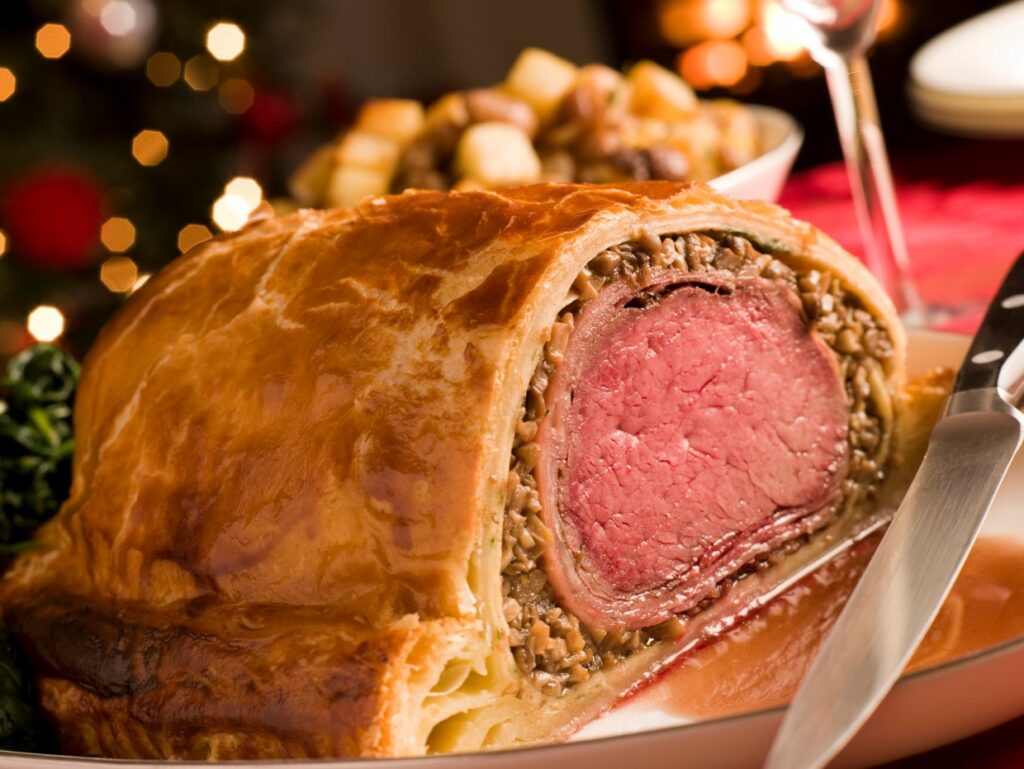 A close-up of a sliced beef Wellington on a plate, showcasing its golden pastry crust and tender beef surrounded by mushroom duxelles. In the background, blurred holiday lights and a bowl of roasted vegetables add a festive ambiance.