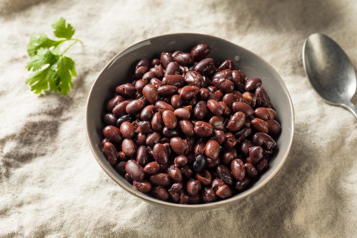 A bowl of cooked black beans on a textured cloth surface. A metal spoon lies nearby, and a sprig of fresh cilantro is in the background. The scene suggests a simple, rustic meal setting.