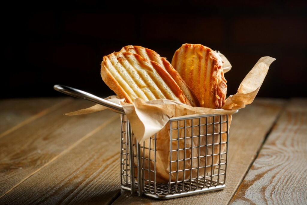 A small metal basket lined with parchment paper holds two golden-brown madeleines on a rustic wooden surface. The background is dark, highlighting the basket and pastries.