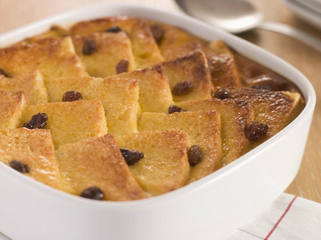 A freshly baked bread pudding in a white square dish, featuring golden-brown, neatly arranged slices topped with raisins. The dish rests on a wooden table, and a spoon is visible in the background.