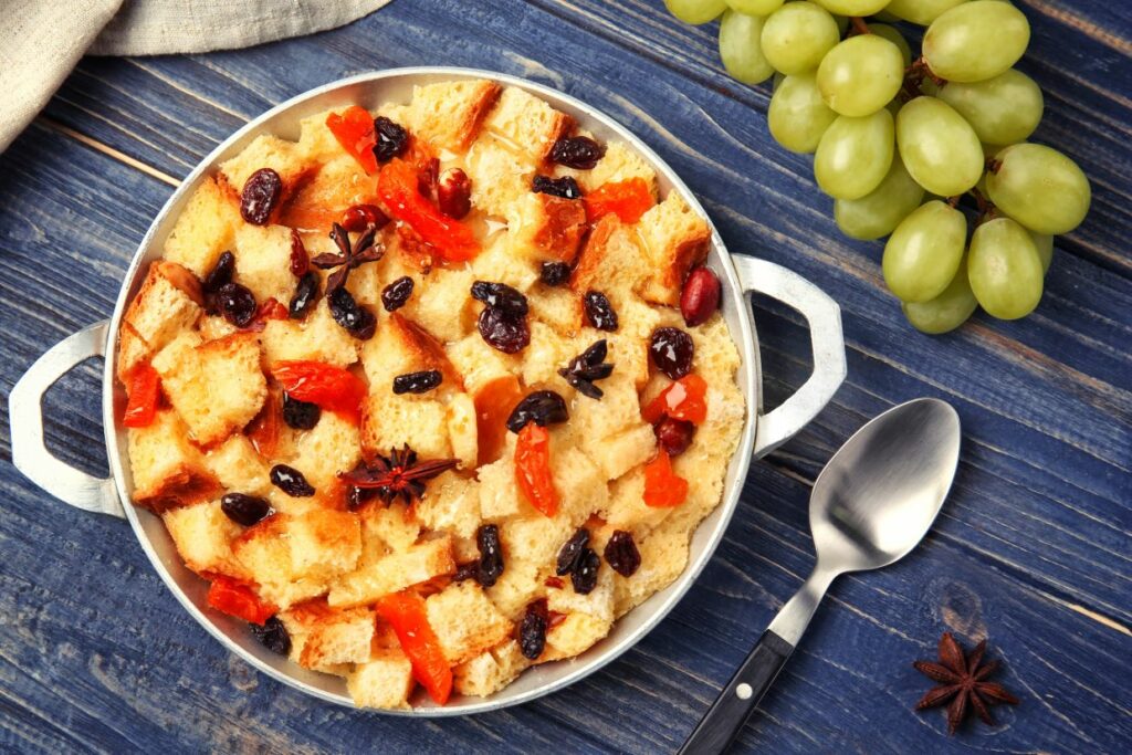 A dish of bread pudding with raisins and red fruit on a wooden table, accompanied by a bunch of green grapes, a spoon, and a star anise.