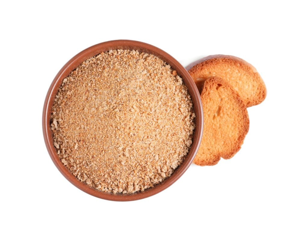 A brown bowl filled with breadcrumbs is placed on a white surface. Next to the bowl are two slices of toasted bread. The breadcrumbs have a fine texture, and the bread slices have a golden-brown color.