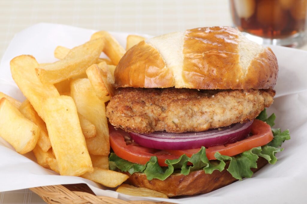 A crispy chicken sandwich with lettuce, tomato, and red onion is served on a pretzel bun, accompanied by a side of thick-cut fries. A glass of iced drink is visible in the background.