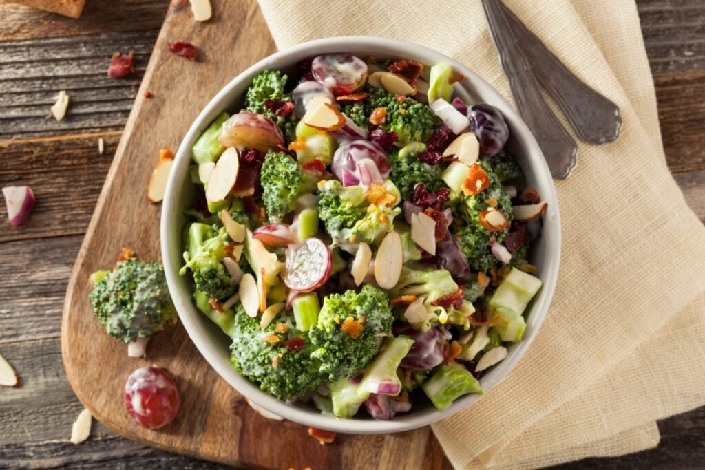 A bowl of broccoli salad on a wooden cutting board, featuring broccoli florets, sliced almonds, red grapes, cranberries, red onion, and a creamy dressing. A beige cloth and a fork are beside the bowl.