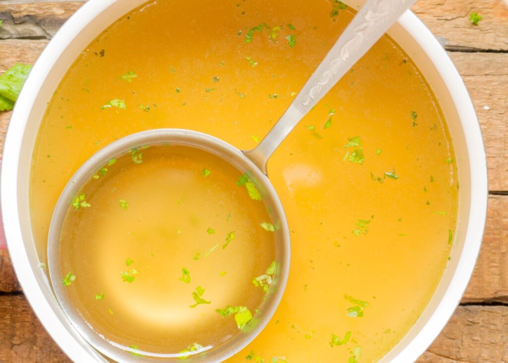 A bowl of clear yellow soup with floating herbs, partially filled and held by a silver ladle. The soup is garnished with chopped green herbs, and the bowl is placed on a wooden surface.