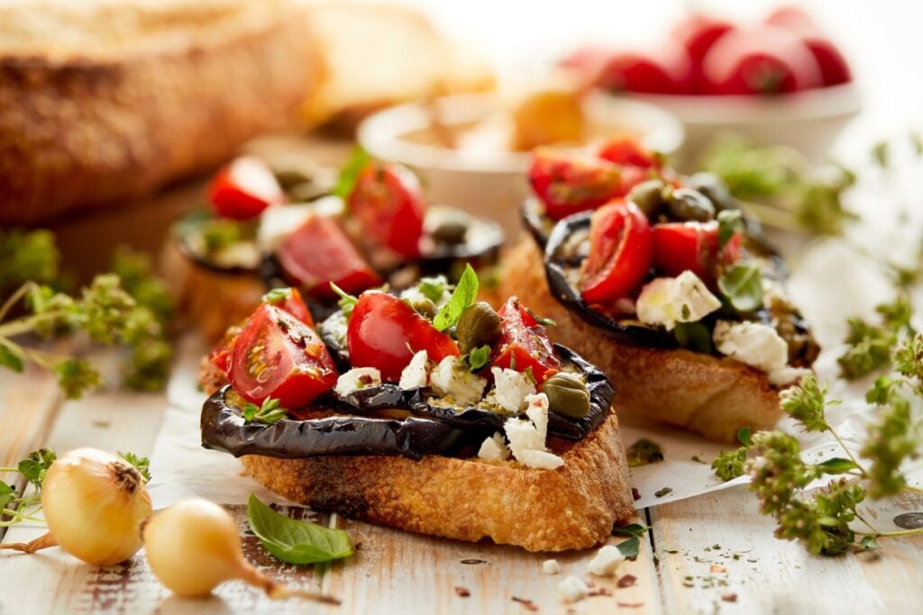 Two slices of toasted bread topped with roasted eggplant, cherry tomatoes, feta cheese, and capers. Garnished with fresh herbs, small onions are scattered around, and a bowl of radishes is in the background on a rustic wooden surface.