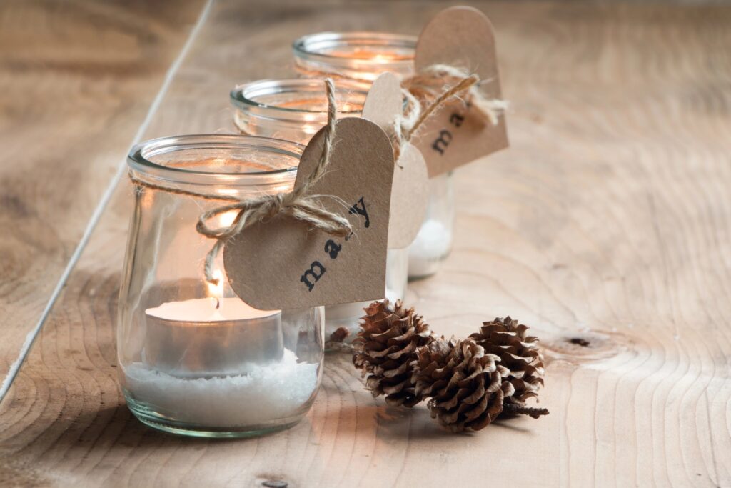 Three glass jars with lit candles inside, each tied with a heart-shaped tag reading "merry," placed on a wooden surface. Snow-like material fills the bottom of the jars, with two pine cones nearby.