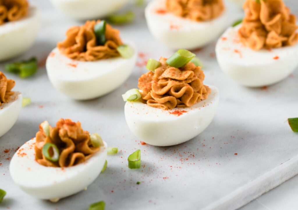 Deviled eggs topped with paprika and sliced green onions are arranged on a light-colored surface. The filling is smooth and creamy, contrasting with the white egg halves. Small onion pieces are scattered around.