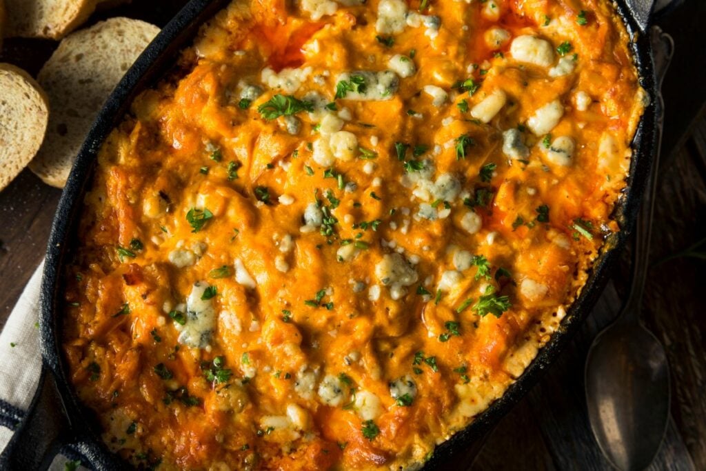 A close-up of a casserole dish filled with a golden, cheesy baked dish. The top is sprinkled with parsley and crumbled cheese. Slices of bread are visible in the background.