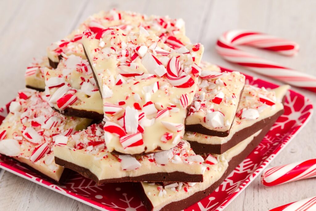 A plate of peppermint bark is stacked, featuring layers of dark and white chocolate topped with crushed candy canes. Several whole candy canes are scattered on the wooden table, adding a festive touch.