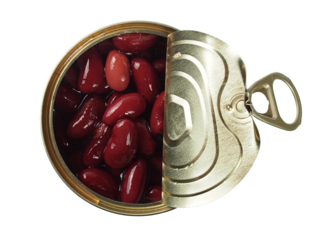 Open can with red kidney beans inside, viewed from above. The lid is partially opened, revealing glossy beans in a light liquid. The can has a metal pull-tab. Bright, high-contrast image on a plain white background.