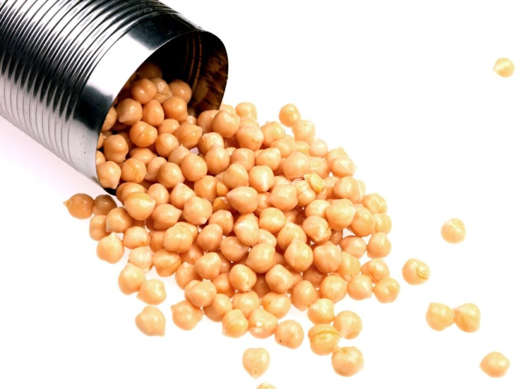 A can tipped on its side, spilling beige chickpeas onto a white surface. The legumes are scattered, showcasing their round shape and smooth texture. The image highlights the simple presentation of canned chickpeas.