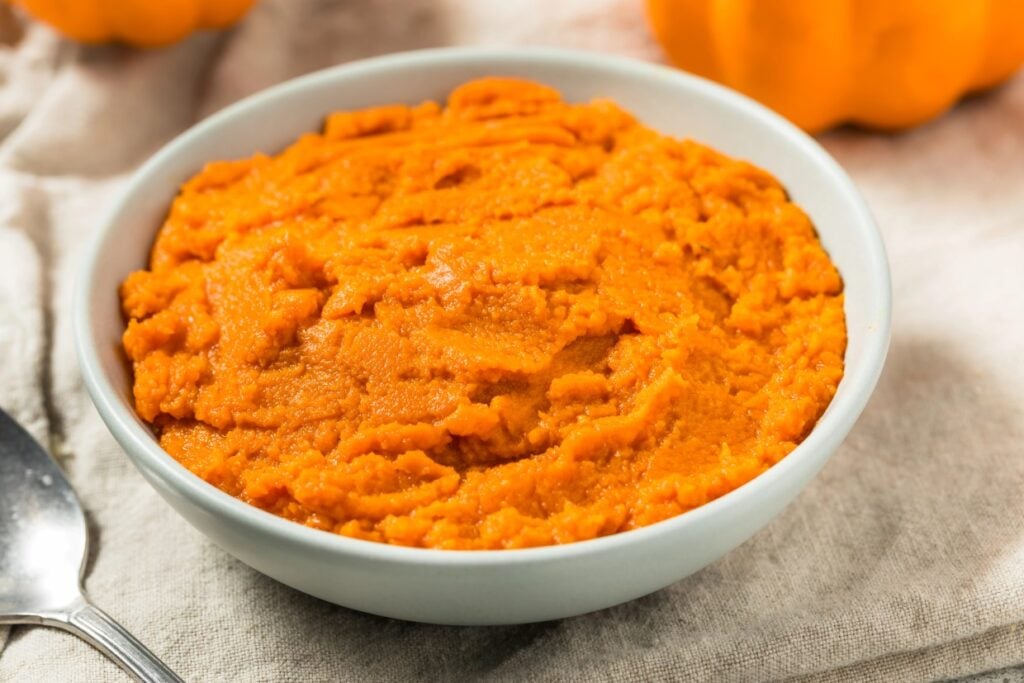 A bowl of smooth, mashed orange pumpkin puree sits on a light-colored cloth. A spoon is placed beside the bowl, and an orange pumpkin can be seen blurred in the background.