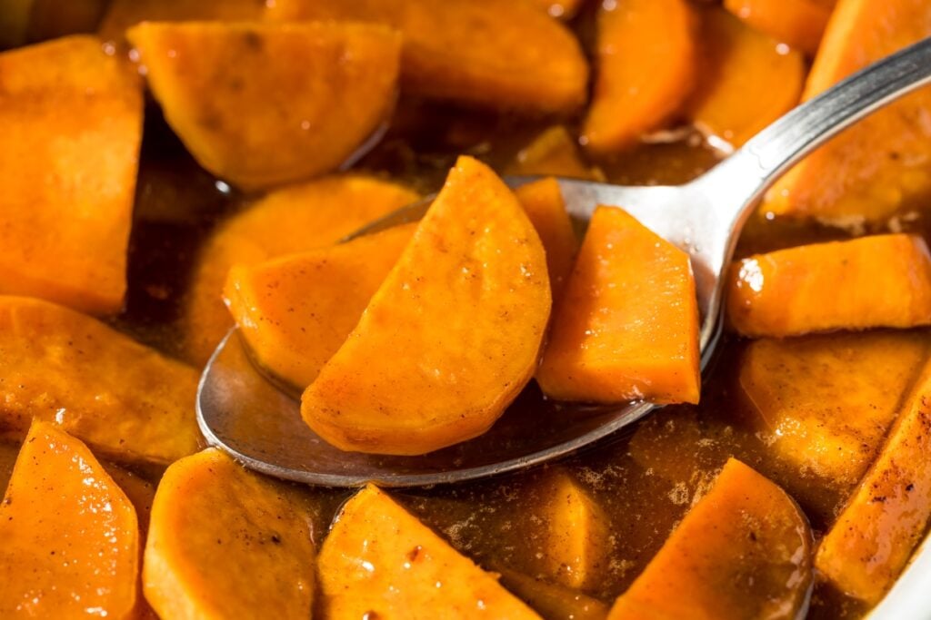 A close-up image of candied yams in a brown syrup. Slices of glazed sweet potatoes are visible, some resting on a spoon, showcasing their glossy, caramelized surface. The warm orange color contrasts with the rich, syrupy sauce.