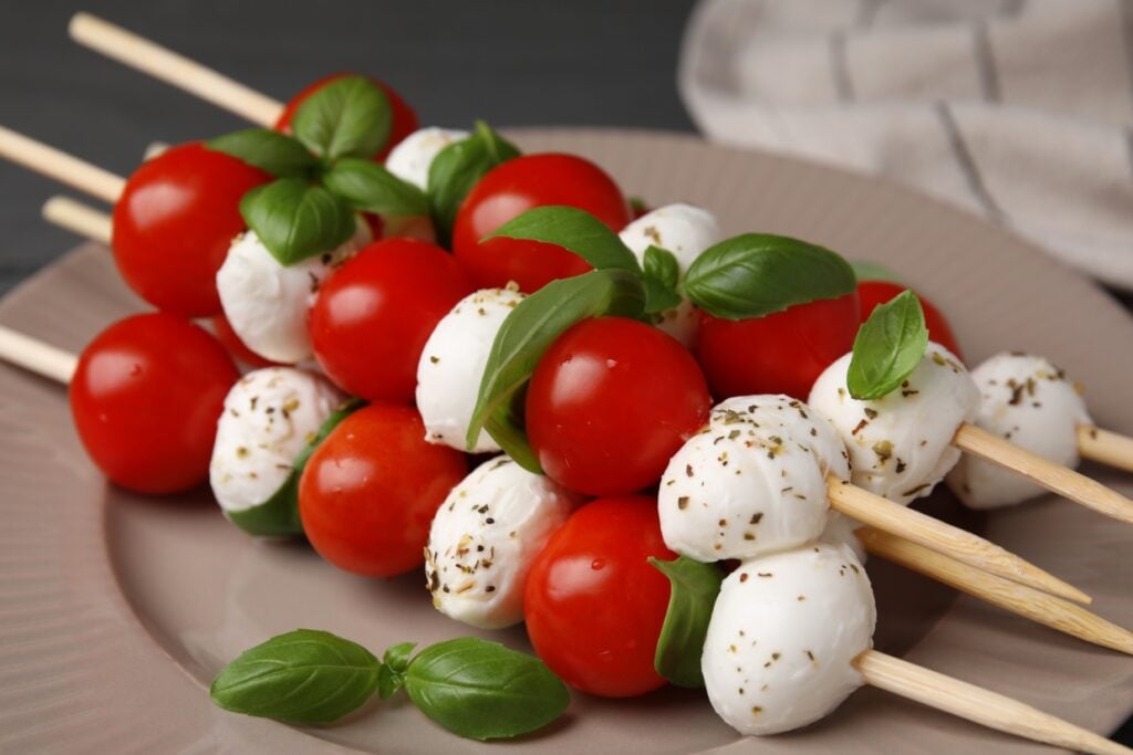 Skewers with cherry tomatoes, fresh mozzarella balls, and basil leaves arranged on a beige plate. The mozzarella is sprinkled with herbs, adding a touch of green and texture. The arrangement is colorful and appetizing.