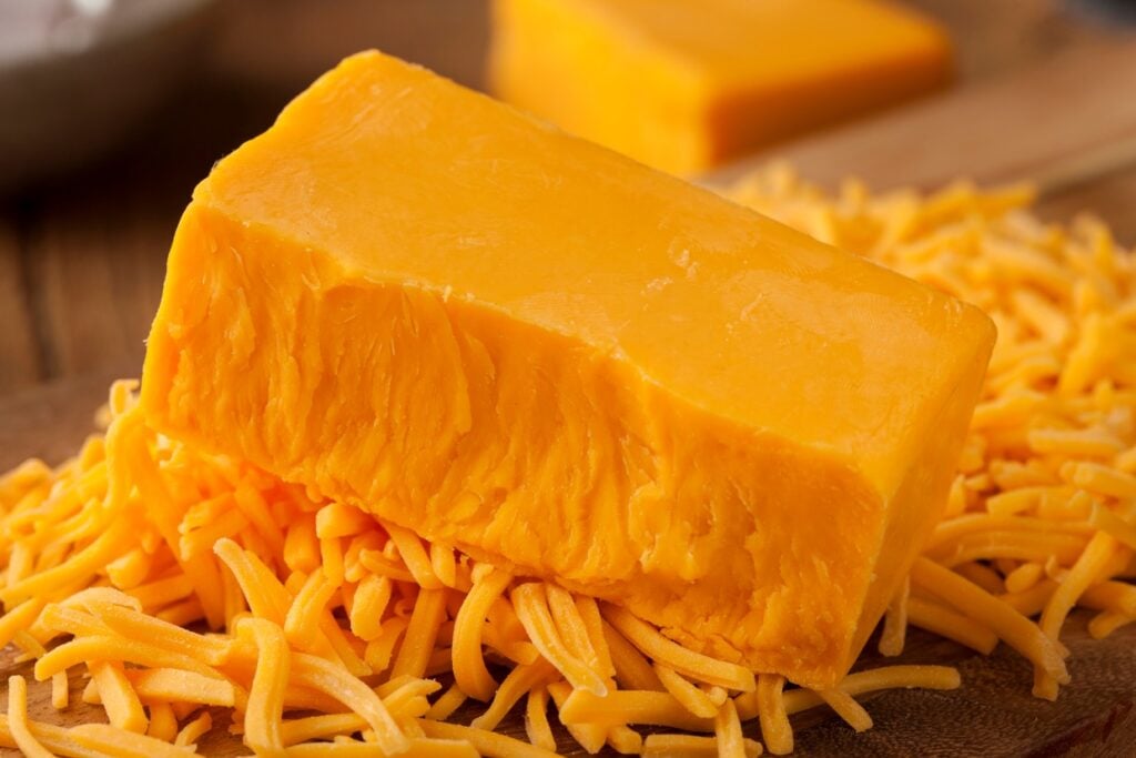 A block of cheddar cheese sits atop a pile of shredded cheddar on a wooden surface. The cheese is a vibrant orange color, with a smooth texture on the block and a slightly rough texture on the shredded pieces.