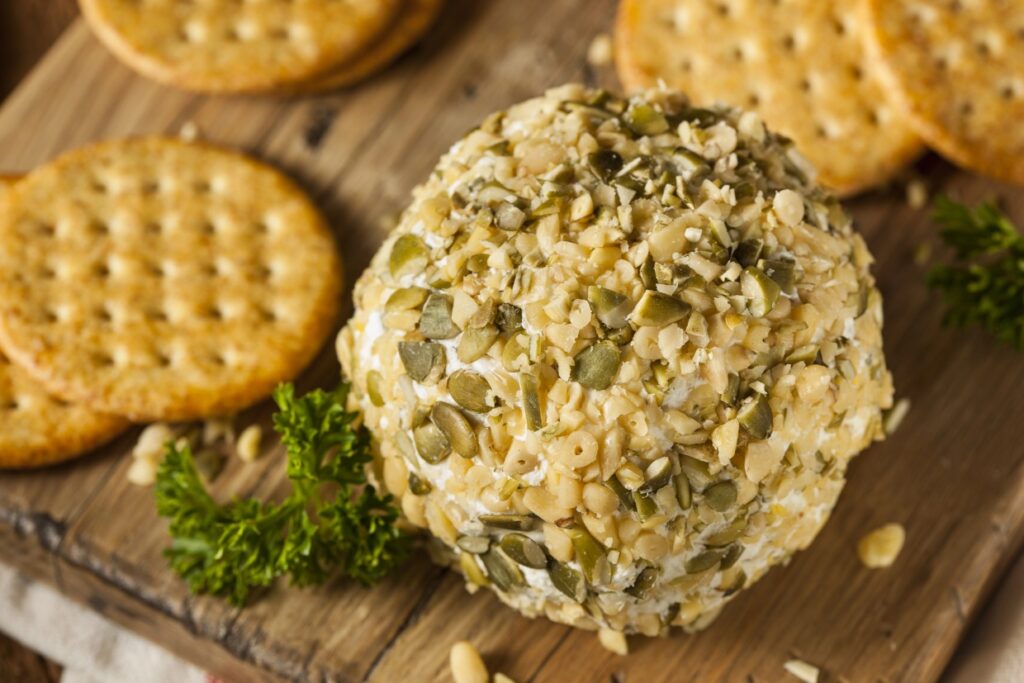 A cheese ball covered in seeds and nuts on a wooden board, surrounded by round crackers. Fresh parsley garnish is nearby, creating an appetizing snack presentation.