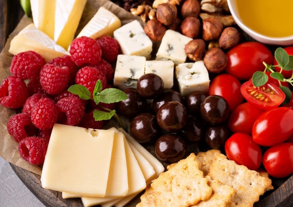 A diverse cheese platter featuring sliced cheese, brie, raspberries, cherry tomatoes, hazelnuts, chocolate-covered treats, blue cheese cubes, crackers, and a small dish of yellow sauce. Mint leaves garnish the raspberries for a fresh touch.