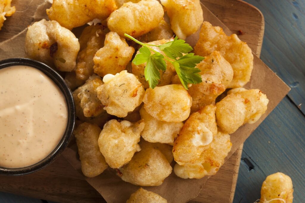 Golden, crispy fried cheese curds are piled on a wooden board with a small bowl of creamy dipping sauce on the side. A sprig of parsley garnishes the top, adding a touch of green.