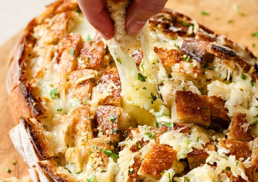 A person's hand pulling a piece of cheesy bread from a loaf filled with melted cheese and herbs. The bread is cut into cubes, displaying a golden-brown crust and gooey cheese. It rests on a wooden surface.