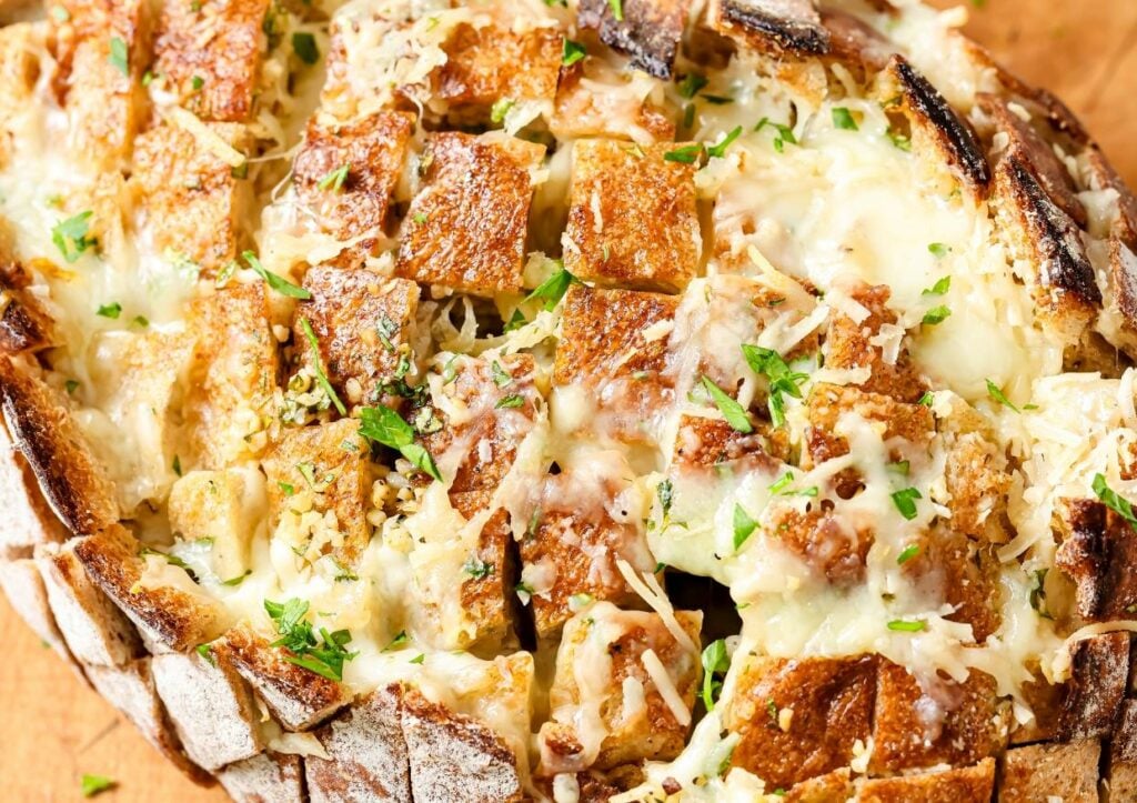 A close-up of cheesy pull-apart bread, with a crusty exterior and melted cheese. The bread is sliced into squares, garnished with chopped parsley, and sprinkled with Parmesan and herbs.