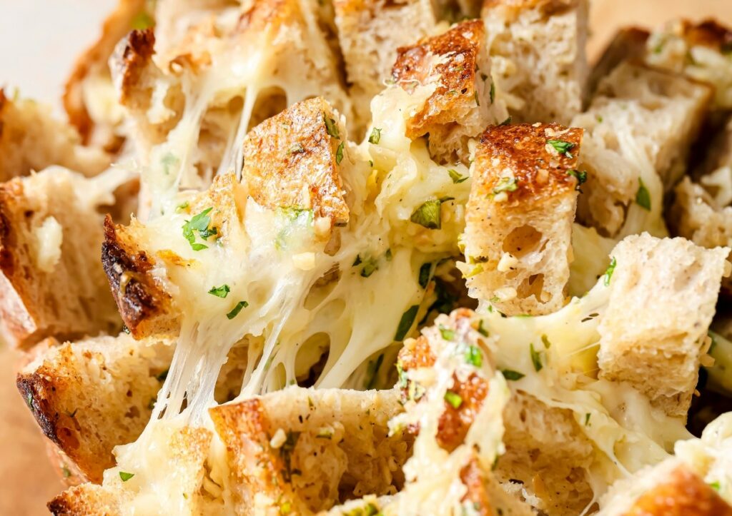 Close-up of cheesy garlic bread with melted cheese pulling between toasted bread cubes, garnished with chopped herbs.