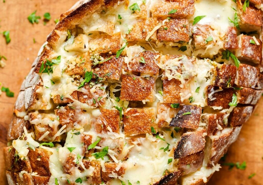 Cheesy pull-apart bread on a wooden surface, garnished with chopped parsley. The bread is sliced in a cross-hatch pattern and filled with melted cheese and herbs.