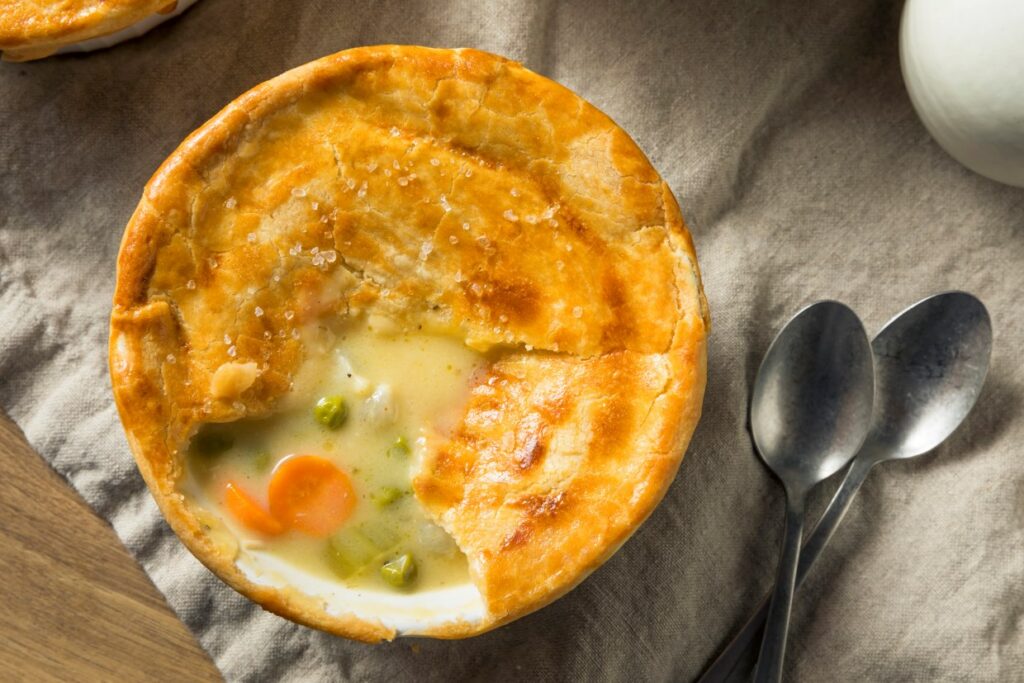 A freshly baked pot pie with a golden crust is partially opened to reveal a creamy filling with peas and carrots. Two spoons rest nearby on a linen cloth.