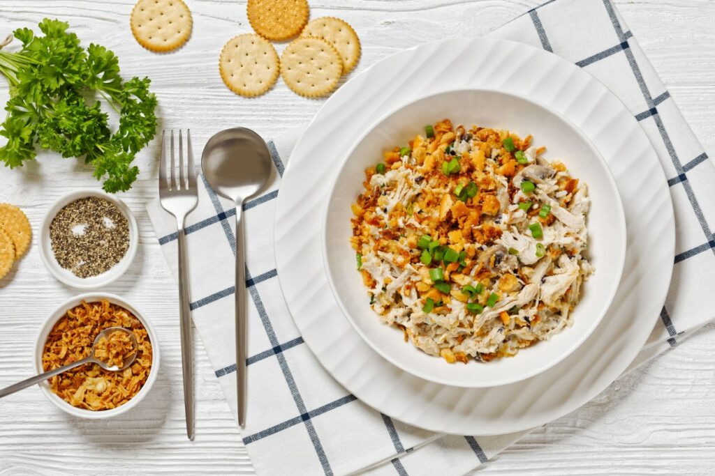 A white plate filled with a dish of shredded chicken, diced vegetables, and croutons, garnished with green onions. Surrounding the plate are crackers, parsley, bowls of fried onions and black pepper, with silver cutlery on a plaid napkin.