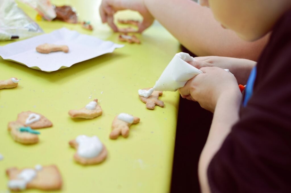 Children decorate animal-shaped cookies with white icing on a green table. Various undecorated cookies and a paper sheet are also visible.