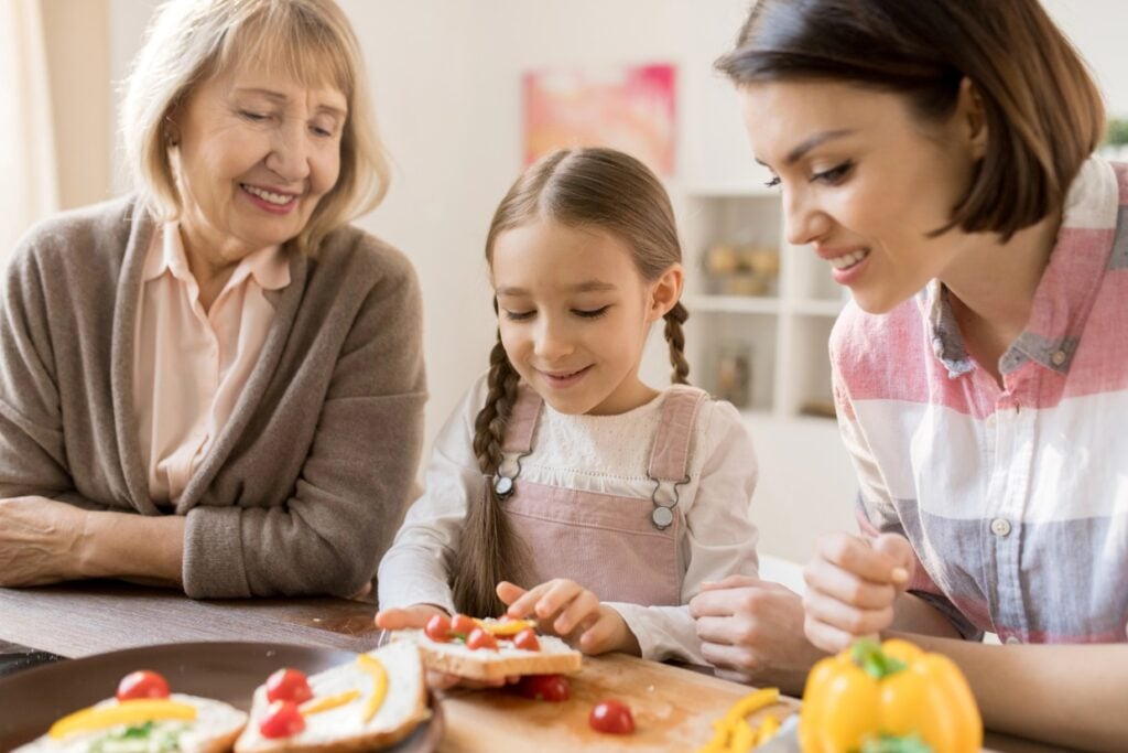 A young girl with braids prepares food with a woman in a plaid shirt and an older woman in a cardigan. They are smiling and working together in a cozy kitchen setting, surrounded by fresh vegetables like tomatoes and a yellow bell pepper.