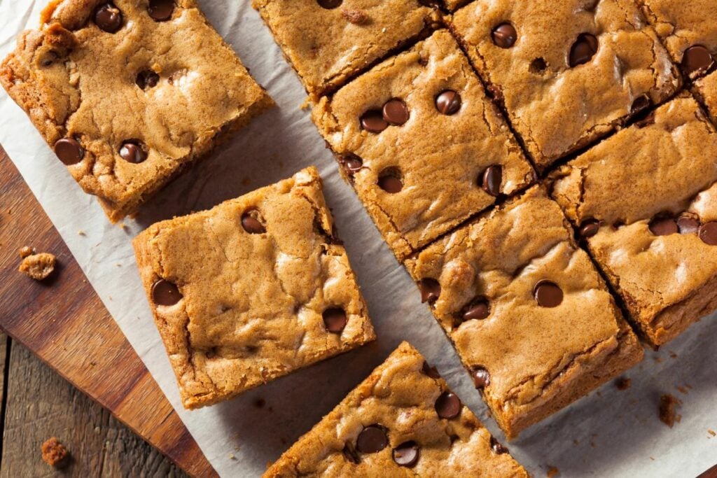 A batch of homemade blondies with chocolate chips, cut into squares on a parchment-lined wooden board. One piece is slightly separated from the others, showcasing the softly baked texture.