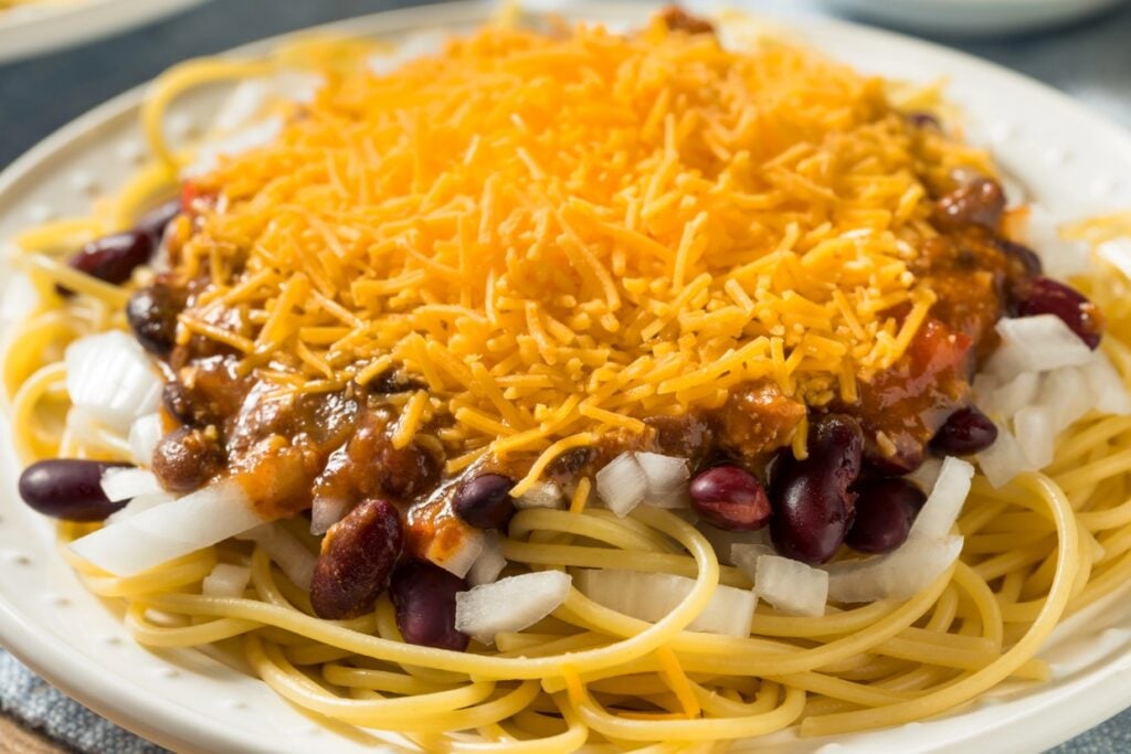 A plate of spaghetti topped with chili, red kidney beans, chopped onions, and a generous layer of shredded cheddar cheese. The dish is served on a white plate.