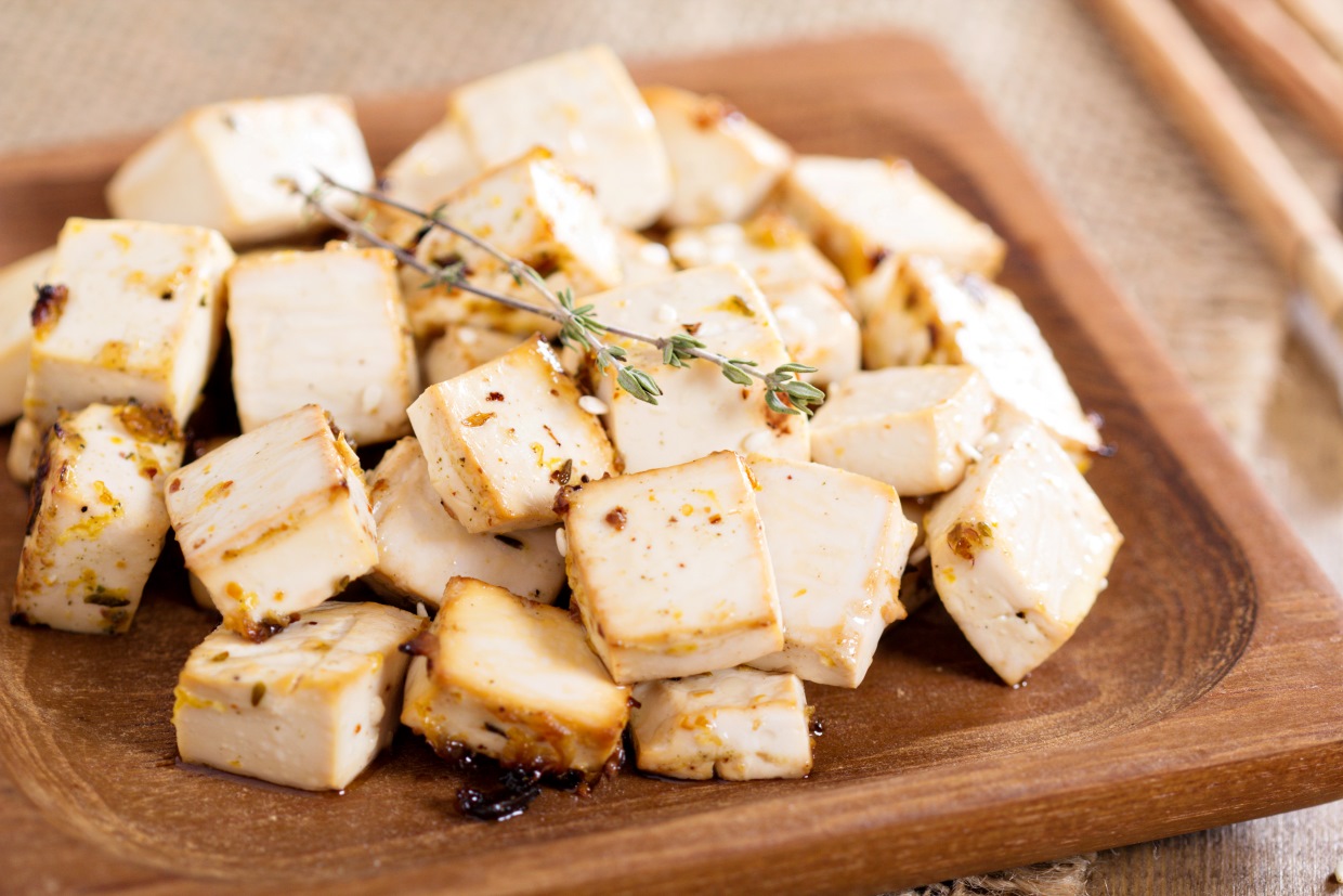 Wooden plate with grilled tofu cubes garnished with herbs. The tofu is golden brown with visible seasoning. The background shows a burlap cloth and a pair of chopsticks.