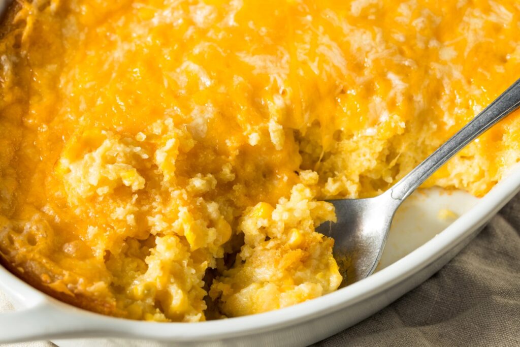 A close-up of a cheesy corn casserole in a white dish. The casserole is golden brown on top with a creamy texture inside, and a serving spoon rests within the dish, revealing a scoop taken out.