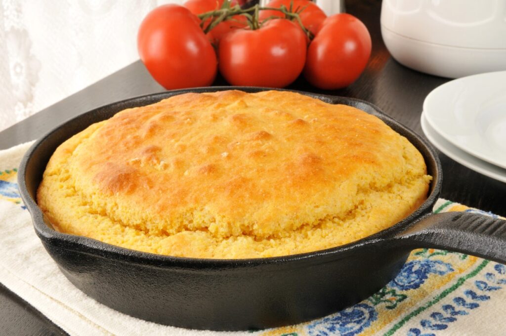 A golden-brown cornbread sits freshly baked in a black cast iron skillet. Three ripe tomatoes are in the background, and a colorful cloth is beneath the skillet.