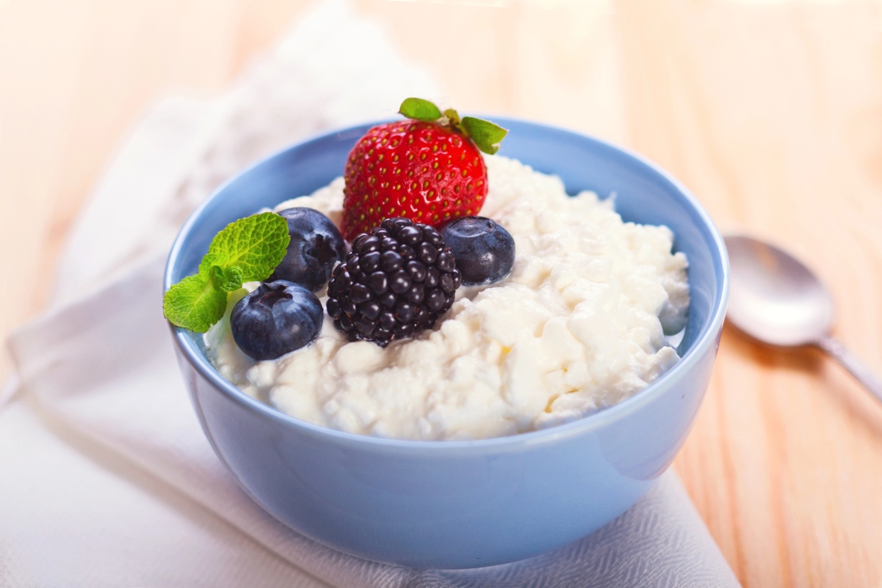 A blue bowl filled with cottage cheese, topped with a strawberry, a blackberry, two blueberries, and a sprig of mint, sits on a wooden table. A folded napkin and a spoon are placed beside the bowl.