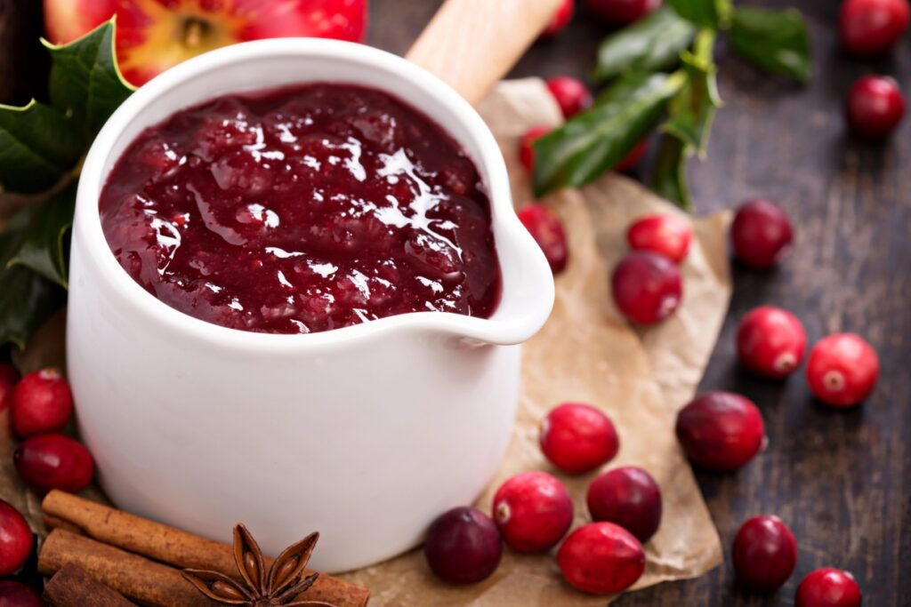 A white bowl filled with cranberry sauce is surrounded by fresh cranberries, cinnamon sticks, and star anise on a wooden surface. Holly leaves and a partially visible red apple are in the background.