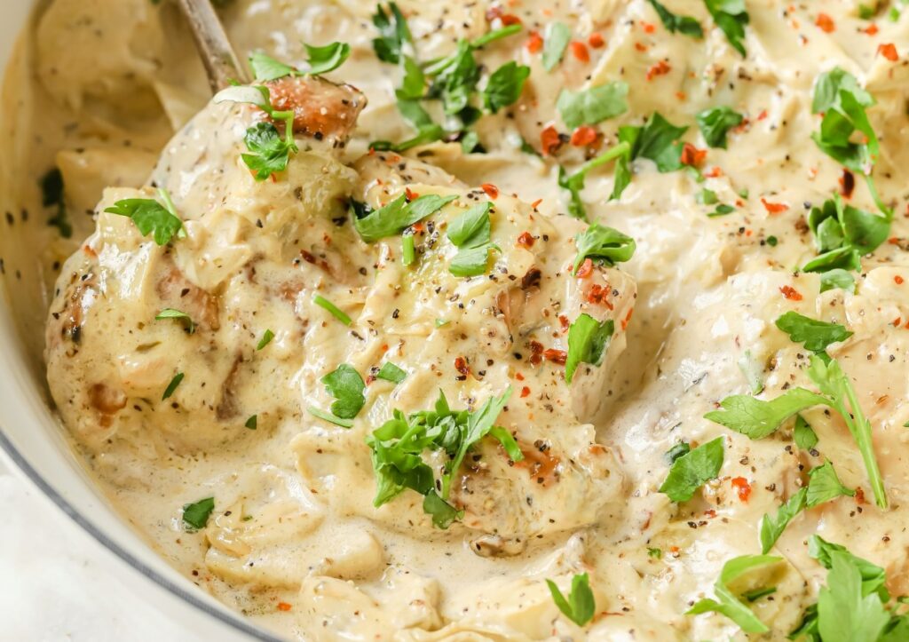 A close-up of a creamy dish featuring chicken pieces in a white sauce, garnished with chopped fresh herbs and red pepper flakes. A spoon is partially visible, suggesting the dish is ready to be served.